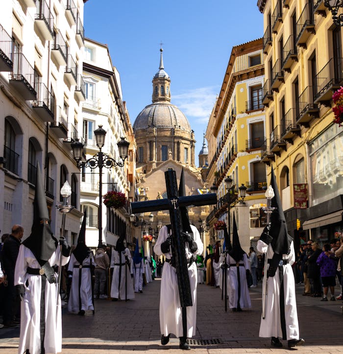Religious procession in Zaragoza with participants in traditional attire and a dome in the background.